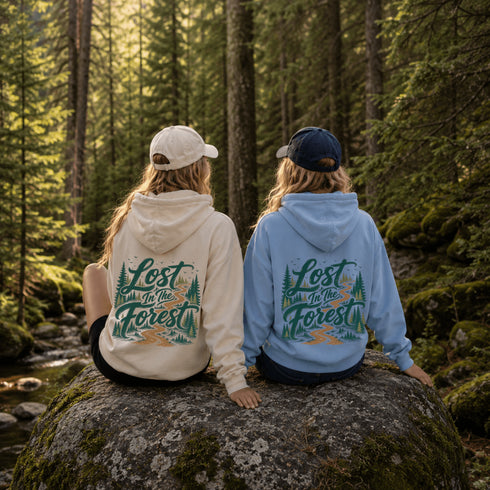 Two people sitting on a mossy rock in a forest, seen from behind, wearing matching hoodies with a large graphic print that reads 'Lost in the Forest.' One hoodie is cream and the other is light blue, both with green and tan artwork.