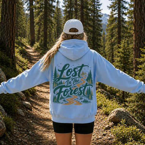 A person standing on a forest trail viewed from behind, wearing a light blue hoodie with a large 'Lost in the Forest' graphic on the back, black shorts, and a white cap.