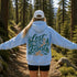 A person standing on a forest trail viewed from behind, wearing a light blue hoodie with a large 'Lost in the Forest' graphic on the back, black shorts, and a white cap.