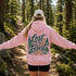 A woman standing on a forest trail viewed from behind, wearing a light pink hoodie with a large graphic on the back that reads “Lost in the Forest” in dark green and tan lettering with pine trees. She is also wearing a white cap, black shorts, and has her arms outstretched.