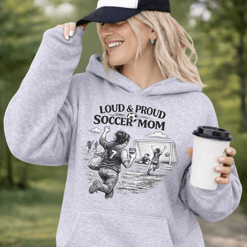 Woman wearing a grey Loud & Proud Soccer Mom hoodie with a vintage soccer scene design, standing on a soccer field with hands in pocket