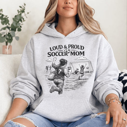 Woman wearing a grey Loud & Proud Soccer Mom hoodie with a vintage soccer scene design, standing on a soccer field with hands in pocket