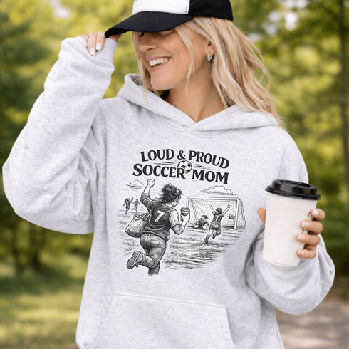 Woman wearing a grey Loud & Proud Soccer Mom hoodie with a vintage soccer scene design, standing on a soccer field with hands in pocket