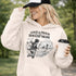 Woman wearing a light tan Loud & Proud Soccer Mom hoodie with a vintage soccer scene design, standing on a soccer field with hands in pocket