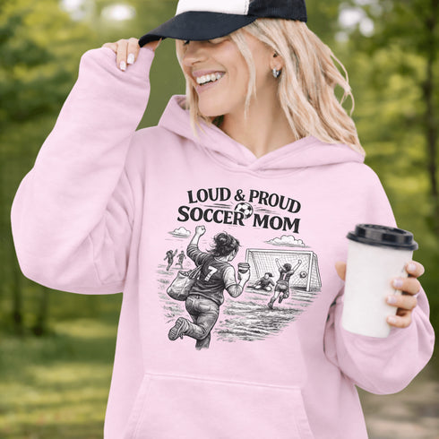 Woman wearing a light pink Loud & Proud Soccer Mom hoodie with a vintage soccer scene design, standing on a soccer field with hands in pocket