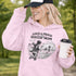 Woman wearing a light pink Loud & Proud Soccer Mom hoodie with a vintage soccer scene design, standing on a soccer field with hands in pocket