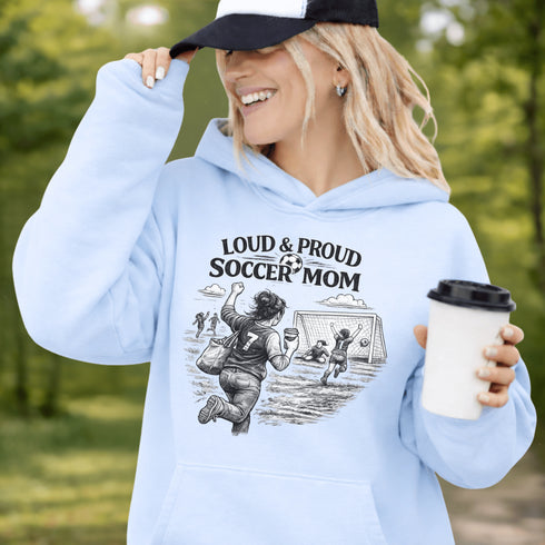 Woman wearing a light blue Loud & Proud Soccer Mom hoodie with a vintage soccer scene design, standing on a soccer field with hands in pocket
