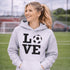 Hoodie with bold LOVE design featuring a soccer ball replacing the O, worn by woman holding coffee cup outdoors