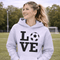 Hoodie with bold LOVE design featuring a soccer ball replacing the O, worn by woman holding coffee cup outdoors