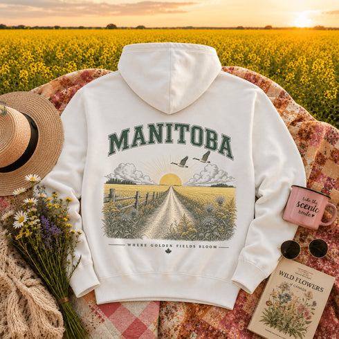 A white pullover hoodie laid flat on a floral blanket in a sunlit field of yellow flowers. The back of the hoodie has a large vintage-style graphic reading "MANITOBA" above a illustrated farm road, fields, clouds, and birds.