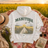 A white pullover hoodie laid flat on a floral blanket in a sunlit field of yellow flowers. The back of the hoodie has a large vintage-style graphic reading "MANITOBA" above a illustrated farm road, fields, clouds, and birds.