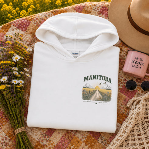 A folded white hoodie with a 'MANIToba' scenic graphic print is styled in a flat lay on a floral quilt, surrounded by a straw hat, sunglasses, a pink candle, and wildflowers.