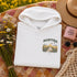 A folded white hoodie with a 'MANIToba' scenic graphic print is styled in a flat lay on a floral quilt, surrounded by a straw hat, sunglasses, a pink candle, and wildflowers.