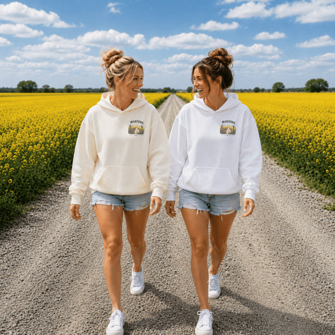 Two women walking hand in hand on a gravel path between bright yellow flower fields under a blue sky. Both are wearing matching white hoodies with a small graphic print on the chest and denim shorts.