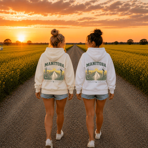 Two women walking away on a gravel road between rows of yellow flowers at sunset, wearing matching white hoodies with a large 'MANITOBA' graphic on the back.