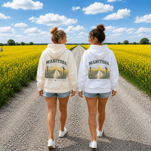 Two women walking away on a gravel path between bright yellow flower fields under a blue sky, wearing matching white hoodies with a large 'MANIToba' graphic on the back.