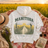 A white pullover hoodie laid flat outdoors on a floral blanket, with a large back graphic reading "MANITOBA" above a rural landscape scene and the phrase "WHERE GOLDEN FIELDS BLOOM." The hoodie is styled with a straw hat, flowers, a pink mug, sunglasses, and a book in front of a golden flower field at sunset.
