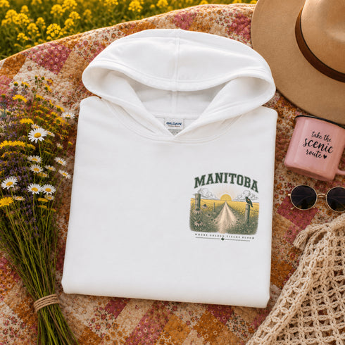 A folded white hoodie with a 'Manitoba' graphic print is styled in a flat lay on a floral blanket with a straw hat, sunglasses, a pink candle, and a bouquet of wildflowers.