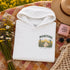 A folded white hoodie with a 'Manitoba' graphic print is styled in a flat lay on a floral blanket with a straw hat, sunglasses, a pink candle, and a bouquet of wildflowers.