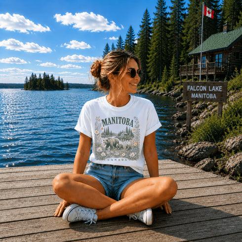 Woman sitting on a wooden dock by a lake with a cabin in the background, wearing a Manitoba t-shirt.