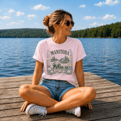 Woman sitting on a dock by a lake wearing a pink 'Manitoba Canada' t-shirt.