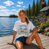 Woman wearing a Manitoba Canada wildlife graphic t-shirt featuring bears, wildflowers, forest landscape and lakeside cabin while sitting on a dock at Falcon Lake Manitoba.