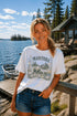 Woman wearing a 'Manitoba Canada' t-shirt by a lake with a cabin in the background