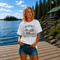 Woman wearing a Manitoba Canada wildlife graphic t-shirt featuring bears, wildflowers, forest landscape and lakeside cabin while standing on a dock at Falcon Lake Manitoba.