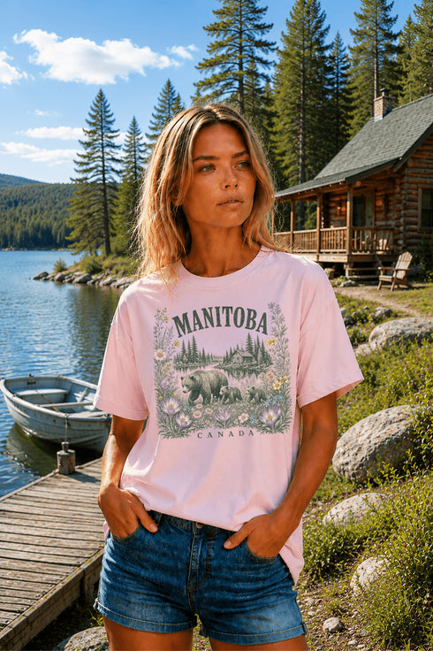 Woman wearing a Manitoba Canada wildlife graphic t-shirt featuring bears, wildflowers, forest landscape and lakeside cabin while standing on a dock at Falcon Lake Manitoba.