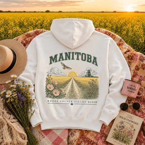 A white hoodie laid flat on a floral blanket with a scenic Manitoba field at sunset in the background. The hoodie has a large back graphic with the word "MANIToba," a flying hawk , a sun, flowers, a grain mill, and the phrase "WHERE GOLDEN FIELDS BLOOM.