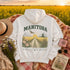 A white hoodie laid flat on a floral blanket with a scenic Manitoba field at sunset in the background. The hoodie has a large back graphic with the word "MANIToba," a flying hawk , a sun, flowers, a grain mill, and the phrase "WHERE GOLDEN FIELDS BLOOM.