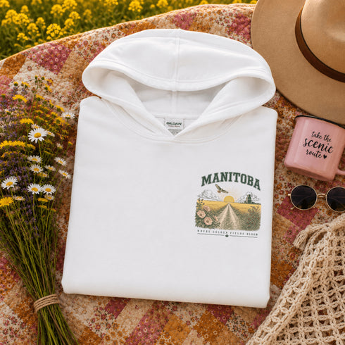 A folded white hoodie with a colorful 'Manitoba' graphic print on the chest, styled in a flat lay on a floral quilt with a straw hat, sunglasses, a pink candle, and wildflowers.