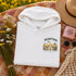 A folded white hoodie with a colorful 'Manitoba' graphic print on the chest, styled in a flat lay on a floral quilt with a straw hat, sunglasses, a pink candle, and wildflowers.