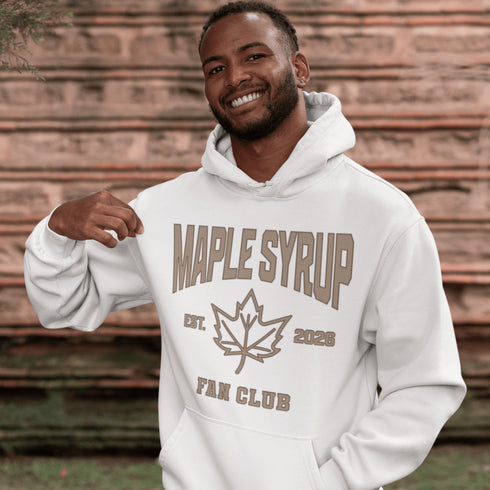 Woman wearing beige Maple Syrup Fan Club hoodie with maple leaf graphic, smiling outdoors in a natural setting with trees in the background