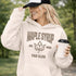 Woman wearing beige Maple Syrup Fan Club hoodie with maple leaf graphic, smiling outdoors in a natural setting with trees in the background