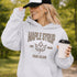 Woman wearing beige Maple Syrup Fan Club hoodie with maple leaf graphic, smiling outdoors in a natural setting with trees in the background