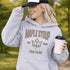Woman wearing beige Maple Syrup Fan Club hoodie with maple leaf graphic, smiling outdoors in a natural setting with trees in the background
