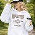 Woman wearing beige Maple Syrup Fan Club hoodie with maple leaf graphic, smiling outdoors in a natural setting with trees in the background