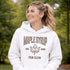 Woman wearing beige Maple Syrup Fan Club hoodie with maple leaf graphic, smiling outdoors in a natural setting with trees in the background