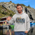 Man wearing a hoodie with a Newfoundland design by a waterfront with boats and cliffs.