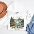 White hoodie with 'Ontario Yours to Discover' design featuring a waterfall, canoe, and blue jay, placed on a white surface with a hat and jeans.