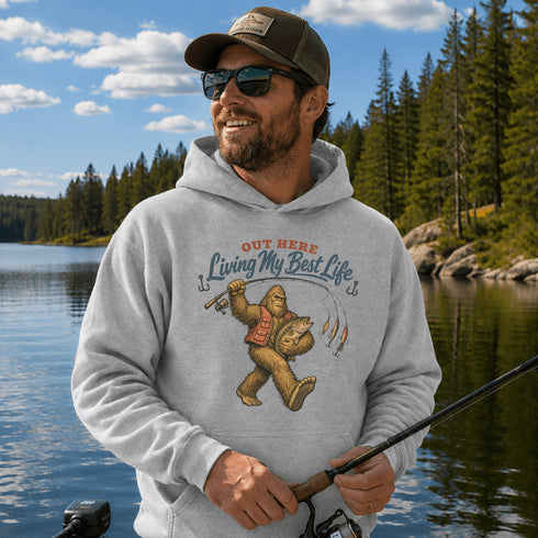 Unisex Gildan 18500 hoodie featuring a vintage-style Bigfoot fishing graphic with the phrase "Out Here Living My Best Life," available in multiple colors.