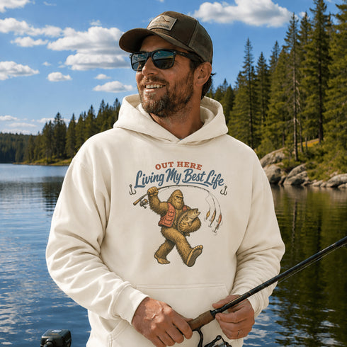 Unisex Gildan 18500 hoodie featuring a vintage-style Bigfoot fishing graphic with the phrase "Out Here Living My Best Life," available in multiple colors.