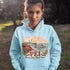 Person wearing a light blue hoodie with a scenic design and 'PEI' text outdoors.