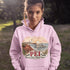 Person wearing a pink hoodie with a scenic design and 'PEI' text outdoors.