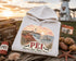 White hoodie with PEI design on a wooden surface with a lighthouse and sunset in the background