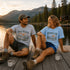 Two people sitting on a dock by a lake with mountains in the background, wearing t-shirts with nature-themed designs.