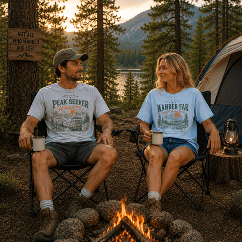 Two people sitting by a campfire in a forest setting, wearing t-shirts with outdoor-themed designs.