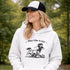 Woman wearing a white Play Like A Girl soccer hoodie with girls soccer graphic, standing on a soccer field with hands in pocket