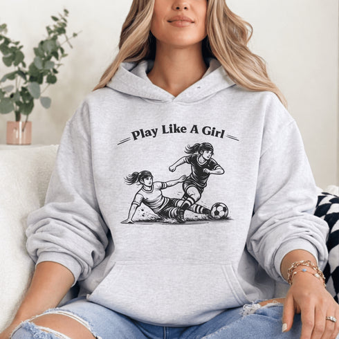 Woman wearing a grey Play Like A Girl soccer hoodie with girls soccer graphic, standing on a soccer field with hands in pocket
